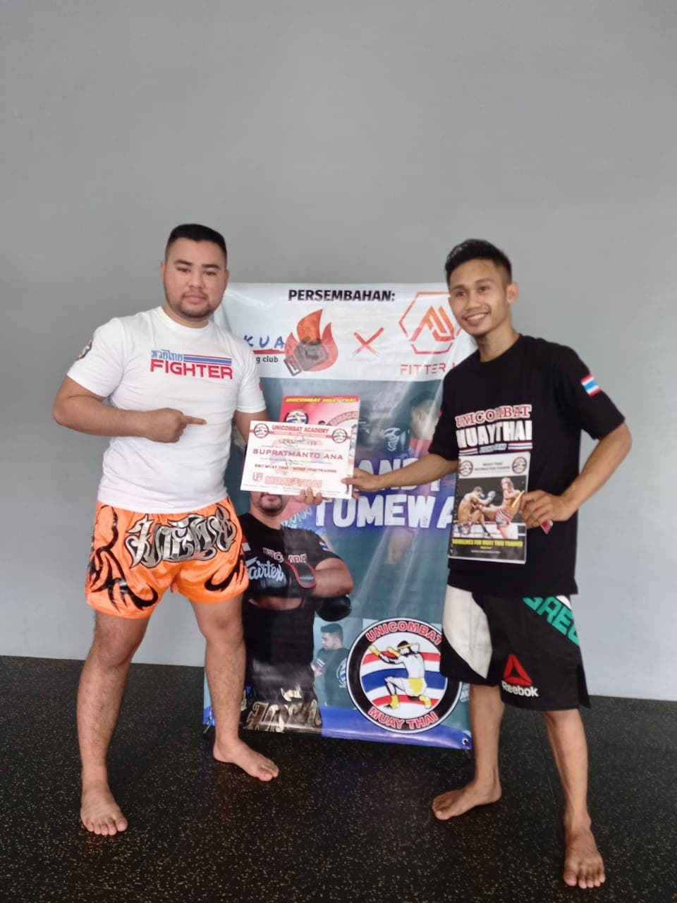 Dua pria berpose memegang sertifikat Unicombat Academy di depan spanduk promosi olahraga Muay Thai.